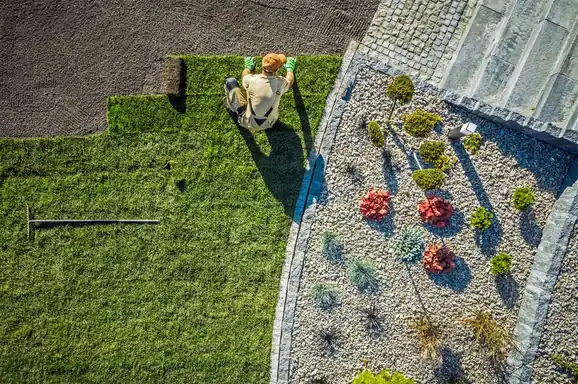 Worker laying fresh sod rolls beside a newly planted garden bed, seen from above