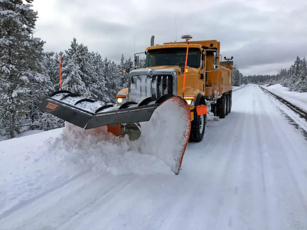 Large orange snow plow truck clearing a snow-covered road lined with snowy trees